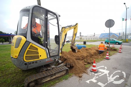 Niewielka koparka kopie wąski rowek na brzegu trawnika, wzdłuż ścieżki rowerowej w pobliżu skrzyżowania ulic. W rowie stoi mężczyzna z łopatą, ubrany w pomarańczową, odblaskową kurtkę. W głębi, lekko zamglone zabudowania. W oddali najbardziej widoczny jest niebieski spadzisty dach.