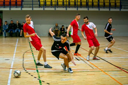 Na parkiecie hali sportowej trwa mecz piłki nożnej halowej. W kadrze trzech zawodników w czerwonych strojach z białą wstawką na lewym ramieniu oraz dwóch zawodników w czarnych strojach z białymi potrójnymi lampasami na ramionach. Jeden z zawodników w czarnym stroju gwałtownie zawraca w stronę piłki leżącej na parkiecie po lewej stronie kadru. Został uchwycony w dynamicznej pozie, pochylony, przykucnięty z rozrzuconymi na boki rękami.