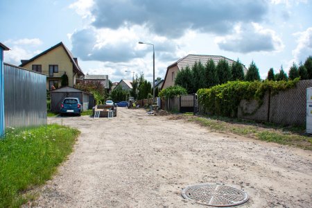 Ulica Andersena przed modernizacją to gruntowa droga. Na wprost budynki jednorodzinne. Po lewej stronie drogi blaszane garaże. Po prawej ogrodzenie, które porasta pnącze.