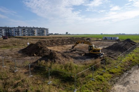 Na pierwszym planie spory obszar ogrodzony siatką, na którym koparka zdejmuje wierzchnią warstwę ziemi i wyrównuje plac. Duży, płytki i płaski wykop otaczają wały ziemi. Po lewej w oddali znajdują się bloki mieszkalne. Po prawej otwarta przestrzeń.