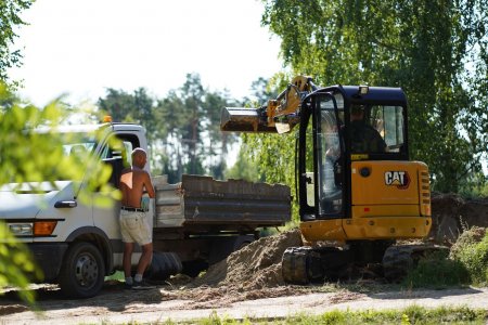 Nieduża ładowarka przesypuje ziemię do stojącej obok wywrotki. Kierowca wywrotki przygląda się pracy ładowarki. Wokół rosną wysokie drzewa.