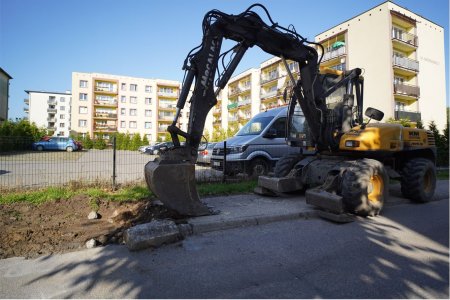 Koparka zrywa stary chodnik i krawężnik. W głębi jasno pomalowane pięciokondygnacyjne bloki, a między nimi parking z zaparkowanymi autami osobowymi i busem.