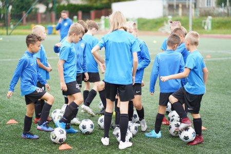 Grupka chłopców w piłkarskich strojach (błękitne koszulki, czarne spodenki i czarne getry do kolan. Każdy z chłopków ma pod nogami piłkę. Stoją na boisku sportowym.