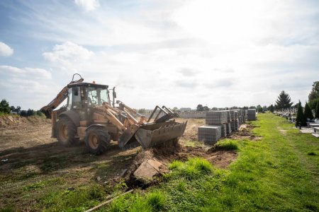 Koparko-ładowarka równa duży plac. Wierzchnia warstwa ziemi z trawą już została zerwana. Na brzegu placu po prawej stronie fotografii leży brukowa kostka w dużych sześciennych paczkach. na skraju po prawej i w oddali na wprost stoją nagrobki cmentarza komunalnego