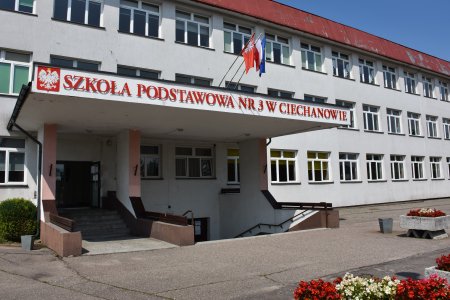 Główne wejście do szkoły podstawowej numer 3. Nad wejściem duże zadaszenie. Na jego krawędzi umieszczono dużymi literami nazwę szkoły oraz flagi: narodową i Unii Europejskiej. Frontowa ściana szkoły jest biała. Widoczny brzeg dachu jest czerwony.