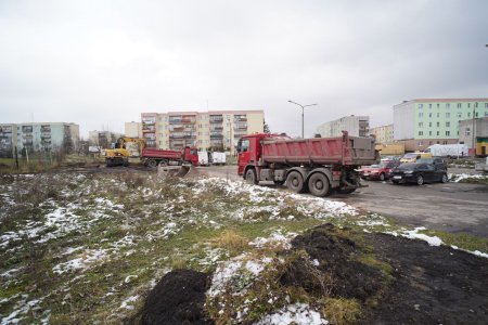 Dwie ciemno czerwone wywrotki i żółta koparka pracują na placu, z którego zrywana jest nierówna, porośnięta chwastami nawierzchnia. Plac otaczają bloki mieszkalne.