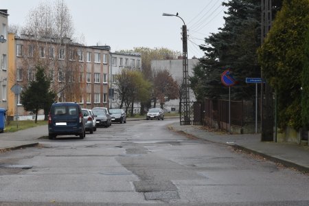 Ulica Asnyka w stanie obecnym, z połataną i nierówną nawierzchnią. Po lewej stronie fotografii znajdują się dwupiętrowe (trzykondygnacyjne) bloki mieszkalne. Na trawniku pod blokami rośnie kilka drzew. Trawnik od jezdni oddziela chodnik. Po lewej drogi stoją auta. Po prawej stronie jezdni biegnie chodnik, a wzdłuż niego znajdują się ogrodzenia posesji, za którymi gęsto rosną wysokie drzewa i krzewy. Na chodniku stoi lampa uliczna, na której szczycie przysiadł ptak.