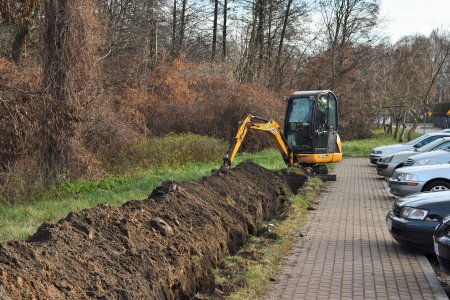 Na zdjęciu nieduża koparka kopie długi dołek wzdłuż chodnika. Po prawej stronie tego chodnika stoją zaparkowane osobowe auta. Po lewej stronie jest wąski pas trawy, dalej są krzewy i wysokie drzewa.