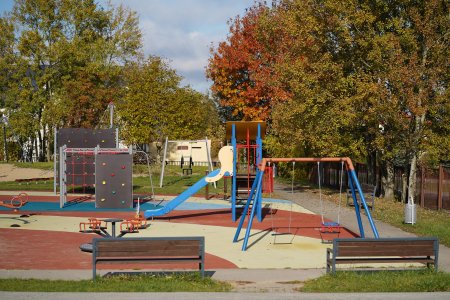 Piękne tło dla placu zabaw stanowią drzewa w jesiennych barwach. Plac zabaw wyposażony huśtawki, zjeżdżalnie i wiele rożnych urządzeń do wspinania.