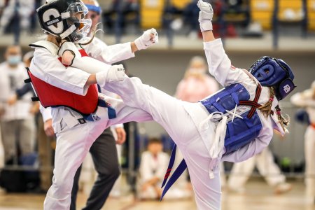 Walka zawodników karate kyokushin. Obaj mają białe stroje i ochraniacze na głowy. Różnią się kolorem kamizelek ochronnych. Zawodnik po lewej ma czerwoną, a po prawej niebieską kamizelkę. Zawodnik po prawej na zdjęciu został uchwycony w czasie wykonywania wykopu. Jego stopa znajduje się pod brodą rywala.