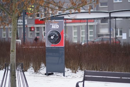 Defibrylator stojący na placu Jana Pawła II (drugiego) za przystankiem autobusowym. Urządzenie jest umieszczone pod przejrzystą kopułą w prostokątnym, pionowym stelażu, oznaczonym literami AED i białym sercem z symbolem błyskawicy.