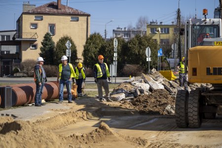 Trwa remont ulicy Asnyka. Na zdjęciu po lewej leżą brązowe karbowane rury o dużej średnicy. Obok rur stoi czterech pracowników w odblaskowych kamizelkach i kaskach. Na środku zdjęcia znajduje się fragment rozkopanego i częściowo wyrównanego piaszczystego podłoża drogi. Po prawej stronie fotografii widać część żółtego pojazdu na dużych podwójnych kołach. W tle zdjęcia jest ulica, a za nią budynki mieszkalne.