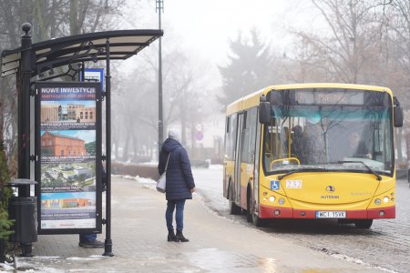 Zdjęcie zrobione w zimowy dzień. Jest biało od śniegu. Po lewej stronie fotografii jest wiata przystankowa, przed którą stoi kobieta. Na przystanek podjeżdża żółto czerwony autobus komunikacji miejskiej (po prawej stronie zdjęcia). Na bocznej ścianie wiaty przystanku znajduje się baner ze zdjęciami dużych miejskich. 