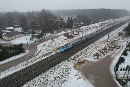 Zdjęcie zrobione z wysoka. Na skos biegną tory kolejowe: od lewego dolnego rogu w górę, w kierunku prawego górnego rogu zdjęcia. Po torach jedzie szybki pociąg Pendolino. Po jednej i drugiej stronie torów jest ulica, która dobiega do torów, dalej skręca i biegnie wzdłuż torowiska. W górnej części zdjęcia jest osiedle domów jednorodzinnych. Rośnie tu dużo wysokich drzew. Na samej górze fotografii znajduje się duży, wysoki las, a nad nim pasek zachmurzonego nieba. Zdjęcie jest biało szare. Wszędzie leży śnieg. 