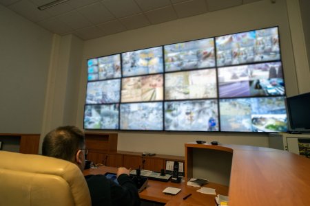 Centrum monitoringu. Na dużej białej ścianie wisi 12 monitorów w trzech rzędach po cztery monitory w rzędzie. Pod monitorami przy ścianie stoi zaokrąglone biurko, przy którym w dużym fotelu siedzi mężczyzna w ciemno niebieskim swetrze.