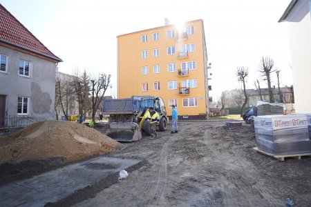 Na zdjęciu znajduje się nieduży plac między budynkami wielorodzinnymi. Po lewej jest fragment bloku mieszkalnego, dalej usypana duża hałda żwiru. Za hałdą stoi spycharka i samochód wywrotka. Obok spycharki znajdują się dwaj pracownicy. Jeden z nich ma jaskrawe ubranie ochronne. Na środku, w głębi zdjęcia stoi pomarańczowy czteropiętrowy blok. Po prawej stronie zdjęcia, pod ścianą jasnego budynku leży dostawa kostki brukowej, owiniętej folią zabezpieczającą.