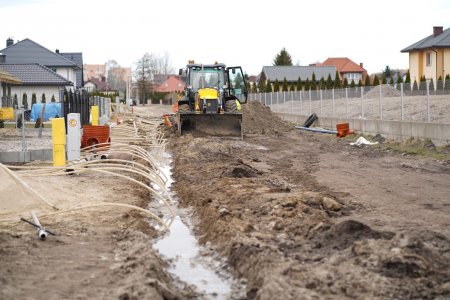 Rozkopana gruntowa droga na osiedlu domów jednorodzinnych. Na środku stoi spychacz. Po lewej stronie drogi wykopany jest niezbyt głęboki rów. Na dnie rowu jest woda. Z rowu, na całej jego długości widocznej na zdjęciu, wystają długie, cienkie rurki, bądź kable, które kładą się na lewą stronę.