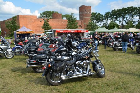 Na błoniach Zamku Książąt Mazowieckich stoją kolorowe namioty. Znajduje się tu również wiele motocykli. W oddali widać zamek i wysokie drzewa. W okolicy namiotów i między motocyklami są ludzie.