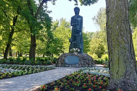 Pomnik Marii Konopnickiej w parku jej imienia. Maria prawą ręką przyciska do piersi książkę, lewą rękę trzyma na lewym ramieniu stojącej przy niej małej dziewczynki. Postaci stoją na postumencie z kamieni, na którym znajduje się owalna tablica. Do pomnika prowadzi chodnik. Po obu stronach chodnika rosną kwiaty. W głębi zdjęcia widać drzewa rosnące w parku.