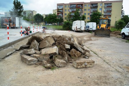 Po lewej stronie zdjęcia jest fragment wcześniej zmodernizowanego parkingu. Na jego skraju stoją biało-czerwone słupki. Ten wyremontowany plac jest wzniesiony wyżej, w stosunku do powstającego, mniej więcej o wysokość stopnia schodów. Na placu budowy na pierwszym planie znajduje się duża gromada starych krawężników i gruzu. Do tej gromady zmierza spycharko-ładowarka, czyli ciągnik z szeroką łychą na przedzie. Po lewej stronie placu pracuje mężczyzna, który tnie beton, wzbijając chmurę białego pyłu. Na obrzeżach stoją trzy białe busy. W głębi jest blok wielorodzinny.