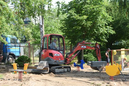 Na przedszkolnym placu zabaw pracuje nieduża koparka. W łysze podnosi kilka betonowych krawężników. Plac zabaw jest rozkopany, gdzieniegdzie stoją bujaki na sprężynach. Za ogrodzeniem przedszkola rosną piękne wysokie drzewa.