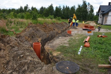 Na środku zdjęcia pionowo biegnie wąski, dość szeroki rów, z którego wystaje duża karbowana pomarańczowa rura. Na dolnym krańcu tego rowka, tuż nad dolną krawędzią zdjęcia, znajduje się studzienka kanalizacyjna zamknięta okrągłą metalową pokrywą. Przy górnym krańcu rowu stoi nieduża żółta koparka, w której siedzi operator. Obok koparki pracuje dwóch mężczyzn w żółtych odblaskowych kamizelkach. Po lewej stronie rowu, za długą hałdą ziemi wykopanej z rowu jest nierówny teren porośnięty chwastami. W prawym górnym rogu zdjęcia znajduje się nowo wybudowany dom, do którego prowadzona jest kanalizacja. Wzdłuż górnej krawędzi zdjęcia jest las.