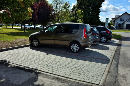 Osobowe auta stoją na nowych miejscach parkingowych. Po lewej stronie stojących samochodów jest teren zielony, na którym rosną duże drzewa. W głębi zdjęcia, za terenem zielonym znajdują się kolejne miejsca parkingowe, zajęte przez samochody.