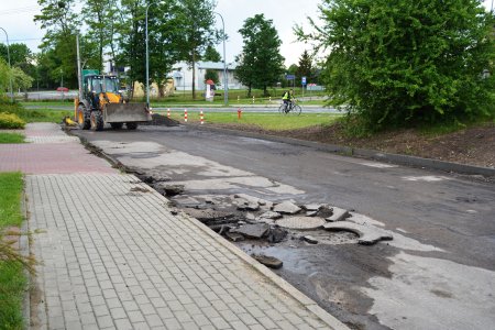 Fragment ulicy Kolbego. W okolicy studzienki kanalizacyjnej leżą kawały pokruszonego asfaltu. Na końcu drogi stoi ciągnik. Po lewej znajduje się chodnik z kostki brukowej. Po prawej rośnie duży krzew. Na wprost widać fragment Alei Niepodległości i rowerzystę.