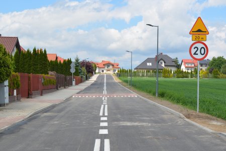 Na środku zdjęcia ulica z białymi liniami, dzielącym pasy ruchu. W połowie drogi wzniesiona wysepka z przejściem dla pieszych. Po lewej jest rząd domów jednorodzinnych, a po prawej pole z zielonymi roślinami. Na poboczu stoi znak z ograniczeniem prędkości do 20 km na godzinę, a nad nim znak ostrzegający o wzniesieniu drogi na wysepce na przejściu dla pieszych za 20 m. Po prawej w oddali i na wprost drogi również są domy jednorodzinne.