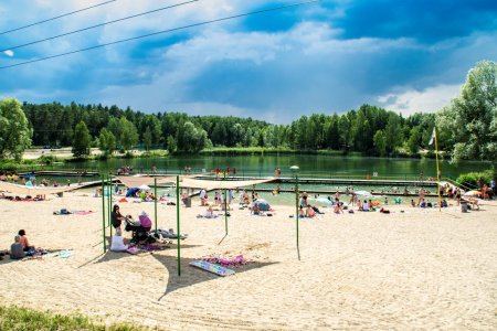 Plaża i zbiornik wodny na Krubinie. Na pierwszym planie, w górnej części plaży, znajdują się żagle przeciwsłoneczne, pod którymi chroni się kilka osób. Dalej, na plaży bliżej wody jest sporo ludzi, w głębi widać pomost na zbiorniku wodnym, otoczonym drzewami.