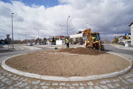 Żółta koparka z szeroką łychą pracuje na nowo wybudowanym rondzie. Koparka przygotowuje grunt, na którym będę zasadzone rośliny. Kolisty środek ronda jest otoczony granitowym krawężnikiem, a na zewnątrz ułożony jest pas granitowej kostki. W głębi zdjęcia, znajduje się szkoła podstawowa.