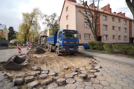 Na środku zdjęcia stoi niebieska wywrotka, a za nią koparka, która przesypuje ziemię. Pojazdy stoją na fragmencie drogi, z której zdjęto dotychczasową nawierzchnię zbudowaną z tak zwanej trylinki, czyli niedużych ale dość grubych, sześciokątnych betonowych bloczków. Po  prawej stronie pojazdów jest dwupiętrowy budynek wielorodzinny. W głębi zdjęcia, po obu stronach drogi znajduje się wiele podobnych budynków, a między nimi drzewa.