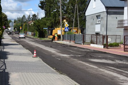 Na zdjęciu po lewej jest chodnik. Po prawej stronie chodnika biegnie ulica, a po prawej stronie ulicy jest rząd prywatnych posesji. Najbliżej jest szary dom za ogrodzeniem, a dalej domy wśród zieleni. Ulica ma zerwaną wierzchnią warstwę asfaltu. Nawierzchnia drogi jest teraz nierówna. W bocznej uliczce po prawej, tuż za szarym domem, stoi wielka żółta frezarka do nawierzchni. Na boku ma napis CAT. Przy maszynie stoi pracownik w odblaskowej kamizelce. W oddali na remontowanej drodze znajdują się dwa samochody osobowe i beczkowóz.