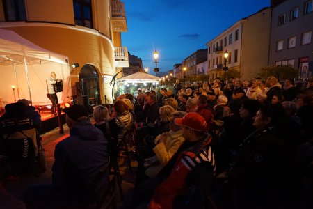 Zdjęcie wykonane po zmroku. Trwa koncert. Przed Kawiarnią W18 tłum ludzi. Na oświetlonej scenie po lewej stoi mężczyzna w białej koszuli. Mężczyzna ma akordeon, zwrócony jest w stronę publiczności. W głębi fotografii widać zadaszony i pięknie oświetlony ogródek dla gości klubokawiarni