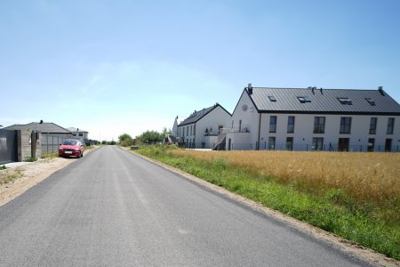 Droga, o równej nawierzchni. Po lewej ogrodzenie prywatnej posesji. Po prawej  dwa podłużne dwukondygnacyjne budynki z balkonami na szczytach.