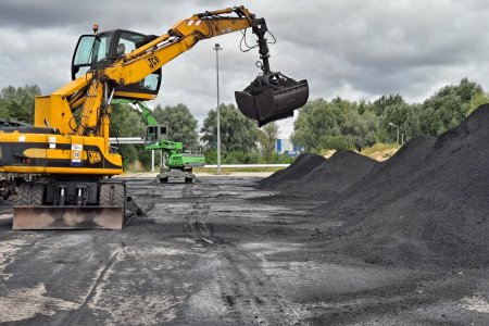 Na dużym wybetonowanym placu pracuje żółta maszyna z długim ramieniem, na końcu którego jest chwytak do przeładowywania między innymi węgla. Maszyna pracuje po lewej stronie, a po prawej jest wielka hałda węgla.