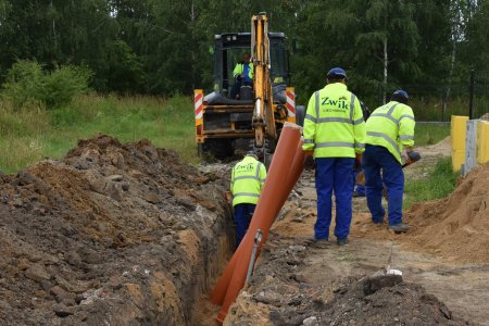 Pracownicy Zakładu Wodociągów i Kanalizacji układają rurę kanalizacyjną w rowie prowadzącym do budynku. Pracownicy mają na sobie jasne, odblaskowe kamizelki.