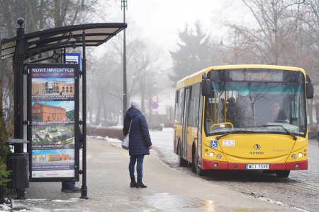 Autobus wjeżdża na przystanek, na którym stoi jedna osoba. Zdjęcie wykonane zimą. Powietrze jest zamglone, gdzieniegdzie leży śnieg. Na bocznej ścianie wiaty przystankowej, za szybą, jest pionowy plakat przedstawiający miejskie inwestycje.