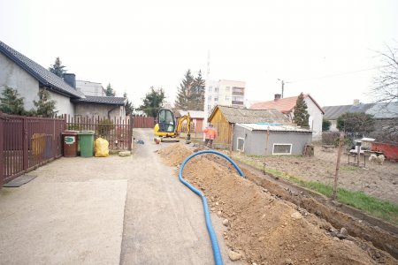 Nieduża żółta koparka kopie długi, niezbyt głęboki rów pod kable oświetleniowe. Fragment kabla znajduje się już w rowie. Kabel jest umieszczony w niebieskiej karbowanej osłonie. Po obu stronach wąskiej uliczki, przy której powstaje oświetlenie znajdują się nieduże prywatne domy. W oddali jest blok wielorodzinny.