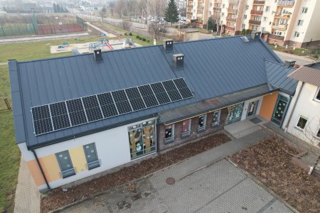 Rząd paneli fotowoltaicznych na dachu podłużnego budynku. Przed budynkiem parking dla aut. W głębi, za budynkiem są boiska i plac zabaw dla dzieci. Wyżej po prawej blok wielorodzinny.