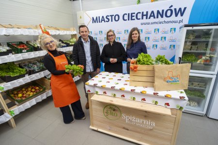 Wnętrze EkoSpiżarni. Na środku, przy ladzie stoi prezydent Ciechanowa Krzysztof Kosiński z dyrektorką MOPS Moniką Lemanowicz oraz dwiema innymi paniami. Jedna z pań ubrana jest w długi pomarańczowy fartuch. Za plecami ludzi ustawiono wielki baner z napisem: Miasto Ciechanów; www.umciechanow.pl i ułożonymi naprzemiennie herbem i logo. Na półkach pod ścianą po lewej znajdują się skrzynki z owocami i warzywami. Produkty żywnościowe znajdują się także w lodówce stojącej po prawej. Na ladzie stoi skrzynka z sałatą, a obok wielorazowa jutowa torba z logo Ciechanowa.