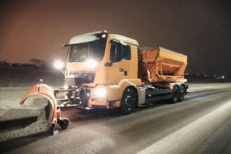 Pomarańczowy pług odśnieża jezdnię. Pracuje nocą, więc drogę oświetlają kierowcy cztery mocne lampy, zamontowane po dwie: na dole i w połowie wysokości kabiny operatora pojazdu.