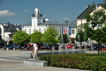 Plac Jana Pawła II latem. Na pierwszym planie fontanna i rabata obsadzona niskimi krzewami. Przy fontannie stoi mężczyzna, który trzyma za rączkę maleńkie dziecko. W głębi placu rosną niezbyt wysokie drzewa o kulistych koronach. Pod drzewami stoją ławki. Wzdłuż jezdni otaczającej plac parkują samochody. Zza drzew wyłania się ciechanowski ratusz o jasnej elewacji.