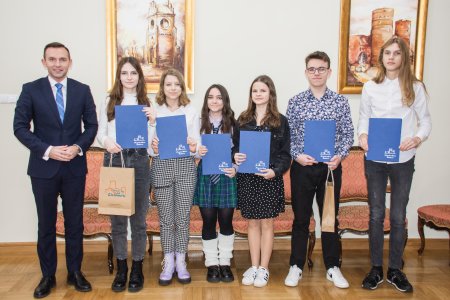 Pamiątkowe zdjęcie młodych stypendystów z prezydentem Ciechanowa Krzysztofem Kosińskim. Zdjęcie wykonano na tle jasnej ściany, na której wiszą dwa obrazy z ciechanowskimi zabytkami: dzwonnicą na Farskiej Górze i Zamkiem Książąt Mazowieckich.