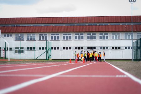 Na pierwszym planie zdjęcia jest nowa bieżnia. W głębi zdjęcia dzieci w kolorowych ubraniach. Tło fotografii stanowi podłużny budynek szkoły - biały z brązowym dachem.