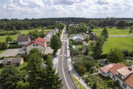 Fotografia wykonana z wysoka, przy użyciu drona. Przez środek zdjęcia pionowo biegnie nowiutka ulica z wyraźnymi liniami dzielącymi pasy ruchu oraz pasami na przejściach dla pieszych. Po obu stronach ulicy znajdują się chodniki oraz posesje z domami jednorodzinnymi. Rośnie tu dużo wysokich drzew. W głębi zdjęcia wzdłuż linii horyzontu rośnie piękny las.