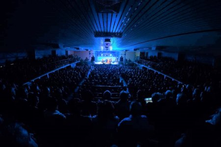 W hali sportowej trwa koncert. W hali jest ciemno. Na niebiesko oświetlona jest tylko scena, znajdująca się daleko w głębi fotografii. Zdjęcie wykonano zza pleców publiczności, oglądającej koncert na balkonie hali.