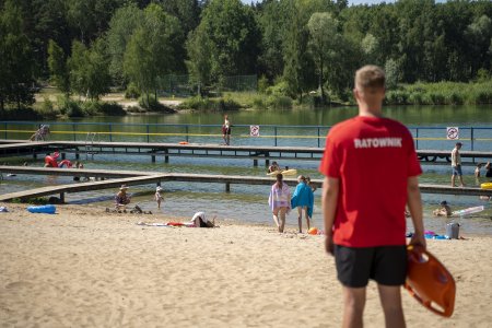 Po prawej stronie zdjęcia stoi ratownik w czerwonej koszulce z bojką ratowniczą w ręku. Mężczyzna stoi na plaży i obserwuje pobliskie kąpielisko. W wodzie kąpie się kilka osób.