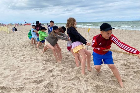 Zdjęcie z wakacji. Na plaży nad morzem spora grupa kolorowo ubranych dzieci bawi się w przeciąganie liny.