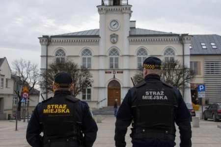 Dwaj miejscy strażnicy w mundurach. Stoją plecami do fotografującego. Na mundurach mają napis: Straż Miejska. Strażnicy idą w stronę ratusza, który jest widoczny w głębi fotografii.